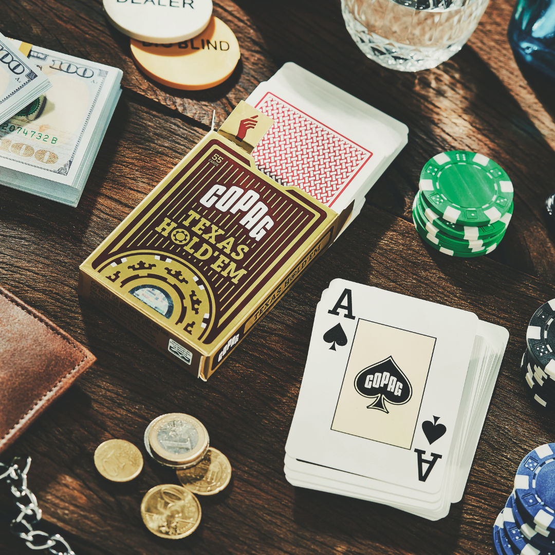 Bildausschnitt einer Tischplatte mit Pokerchips, Geldscheinen und -münzen sowie einer Brieftasche, einem Glas und im Mittelpunkt einer geöffneten Schachtel Copag Poker Karten Texas Hold'em mit oben herausgeschobenen Spielkarten von der Rückseite und einem Kartenstapel mit der Frontseite des Pik Asses.