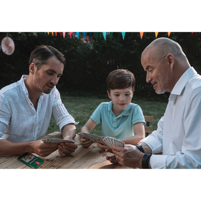 Ein Großvater, Sohn und Enkel spielen im Garten an einem Holztisch mit den ASS Altenburger Doppelkopf-Spielkarten.