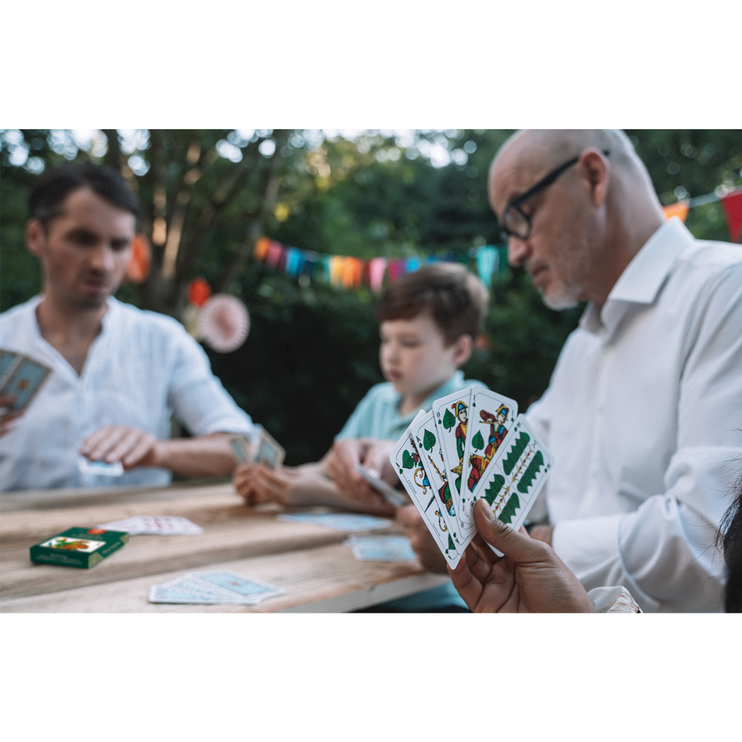 Ein Großvater, Sohn und Enkel spielen im Garten an einem Holztisch mit den ASS Altenburger Schafkopf/Tarock Spielkarten.
