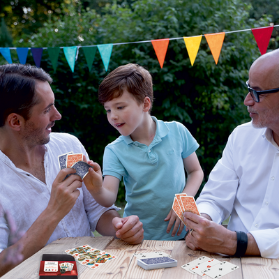 Ein Großvater, Sohn und Enkel spielen im Garten an einem Holztisch mit den ASS Altenburg Senioren-Rommé Spielkarten.