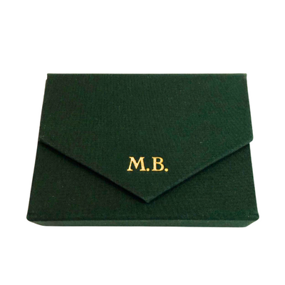 Ein geschlossenes dunkelgrünes Spielkarten-Etui bezogen mit dunkelgrünem Stoff mit einem  goldgeprägten Monogramm M.B. auf der Verschlusslasche.