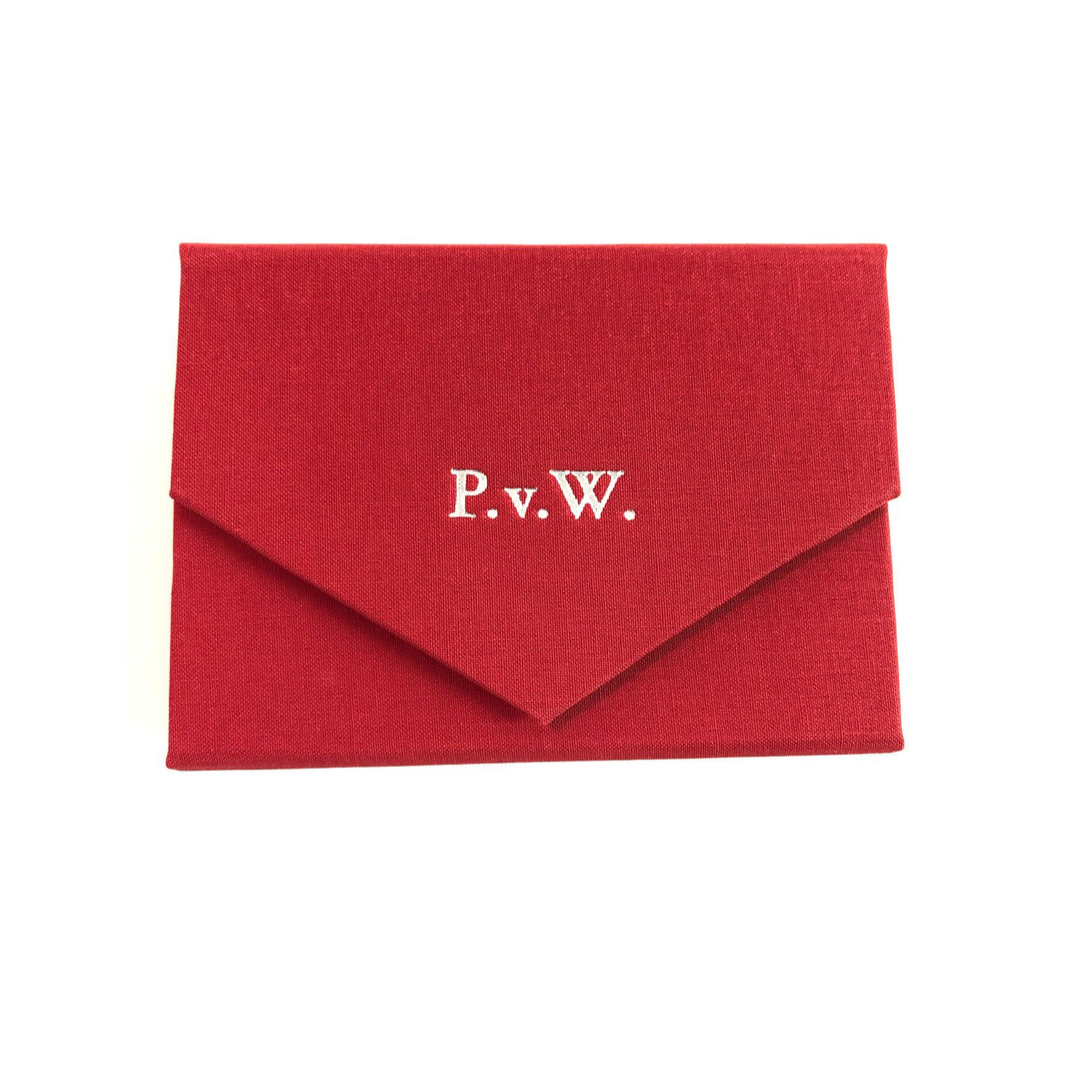 Ein geschlossenes Spielkarten-Etui bezogen mit rotem Stoff mit einem  silber geprägten Monogramm P.v.W. auf der Verschlusslasche.