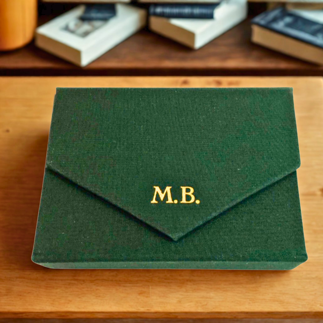 Ein geschlossenes dunkelgrünes Spielkarten-Etui bezogen mit dunkelgrünem Stoff mit einem  goldgeprägten Monogramm M.B. auf der Verschlusslasche auf einem Holzregal.